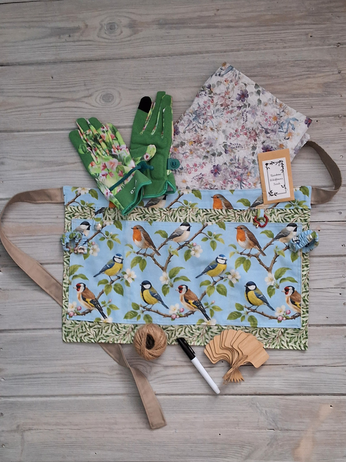 A birds-themed fabric gardening apron with a garden birds illustration print, accompanied by ladies green floral gardening gloves, embedded seed paper, jute twine, bamboo plant labels, and pen, all arranged on a gray wooden plank background