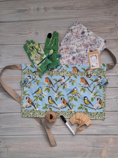 A birds-themed fabric gardening apron with a garden birds illustration print, accompanied by ladies green floral gardening gloves, embedded seed paper, jute twine, bamboo plant labels, and pen, all arranged on a gray wooden plank background