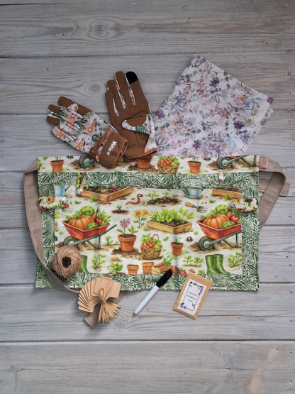 A gardening-themed fabric gardening apron with a vintage illustration print, accompanied by ladies beige tan daisy gardening gloves, embedded seed paper, jute twine, bamboo plant labels, and pen, all arranged on a gray wooden plank background.
