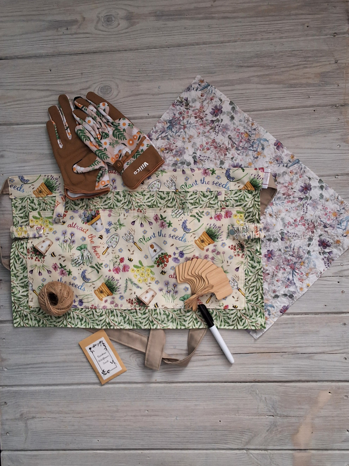A planting-themed fabric gardening apron with a vintage seeds and bee print, accompanied by ladies beige tan daisy gardening gloves, embedded seed paper, jute twine, bamboo plant labels, and pen, all arranged on a gray wooden plank background