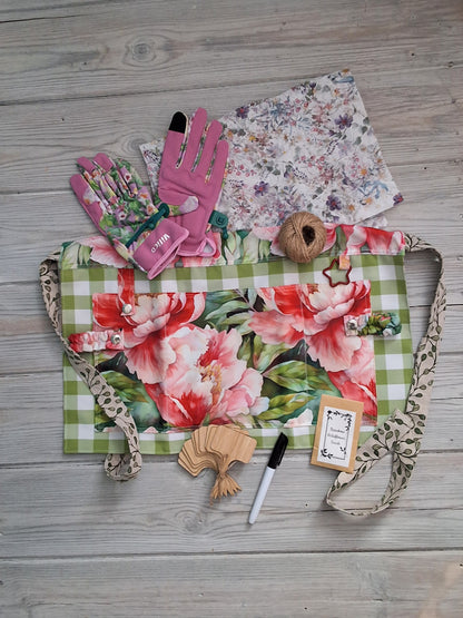 A peony and rose-themed fabric gardening apron with a vintage illustration print, accompanied by ladies pink floral fabric gardening gloves, embedded seed paper, jute twine, bamboo plant labels, and pen, on green checkered background 