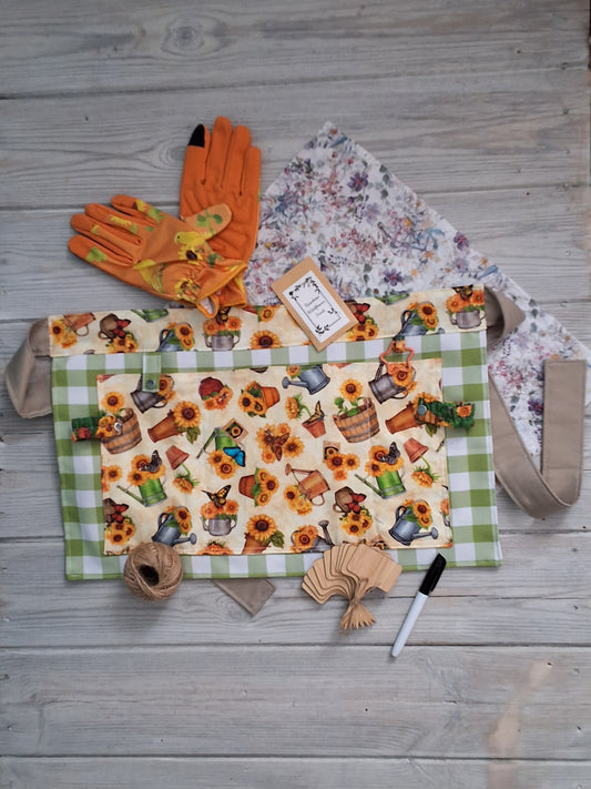 A sunflowers and watering cans-themed fabric gardening apron with a vintage illustration print, accompanied by orange fabric ladies gardening gloves, embedded seed paper, jute twine, bamboo plant labels, and pen, green checkered background 