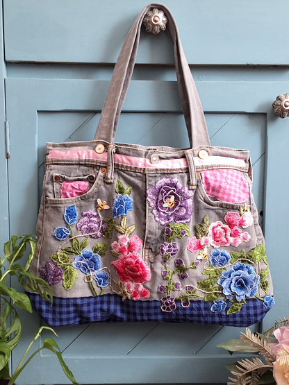 A denim upcycled handmade tote bag front view. With colorful floral embroidery, pink gingham pockets, and a blue gingham bottom trim, hanging against a light blue paneled door.